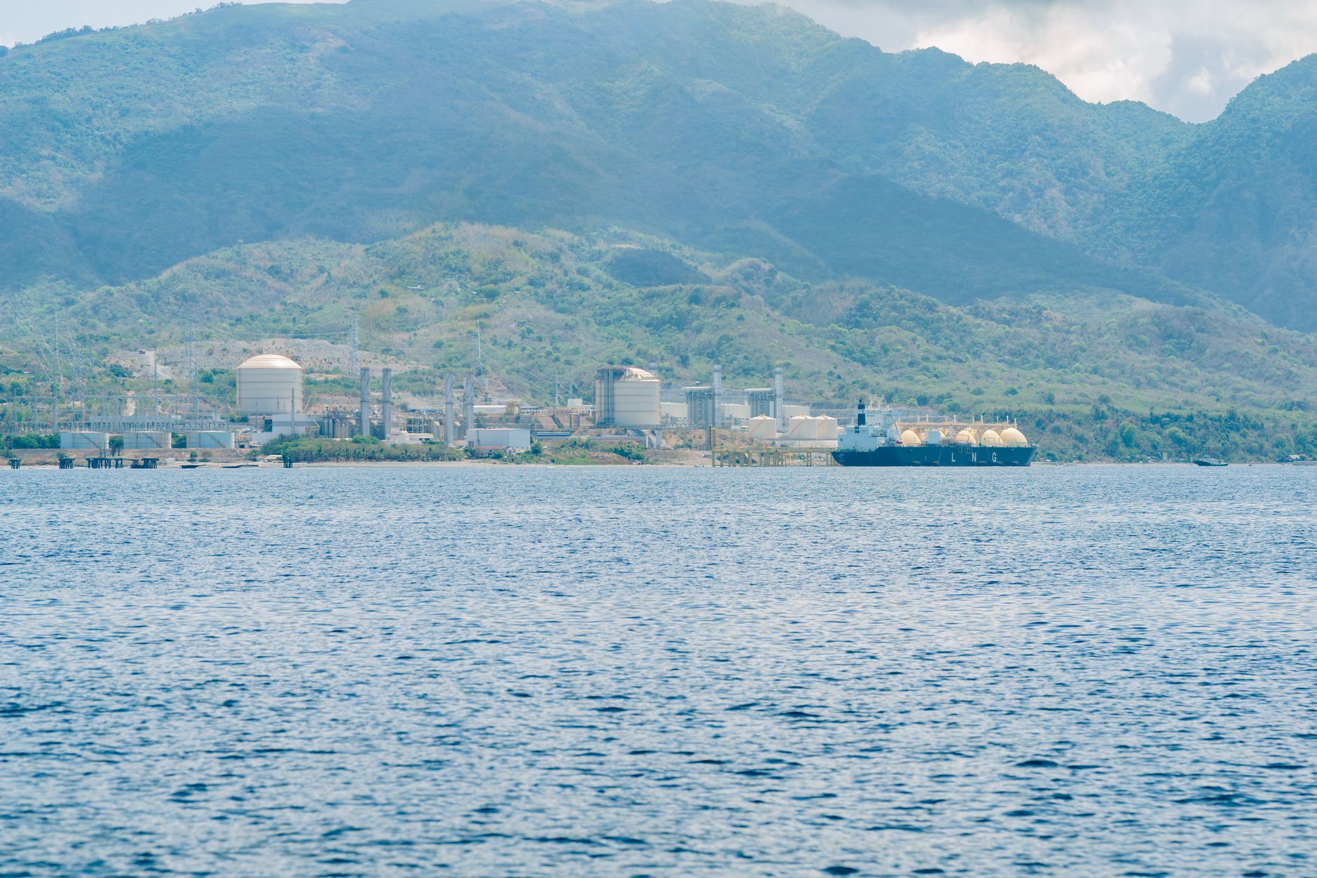Industrial facility by the coast with a docked LNG tanker, set against mountain backdrop.