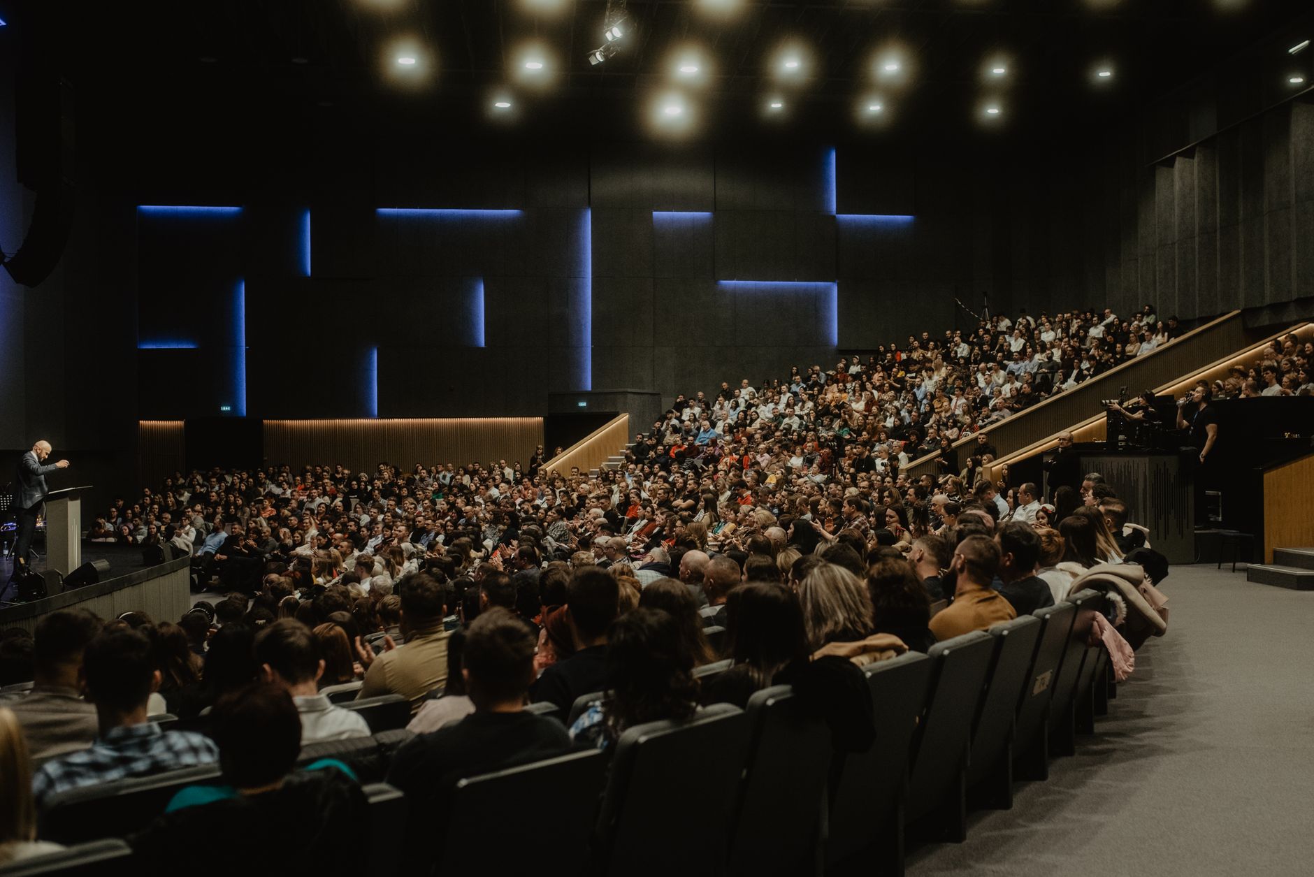 A large audience listens to a speaker at a podium in a conference hall