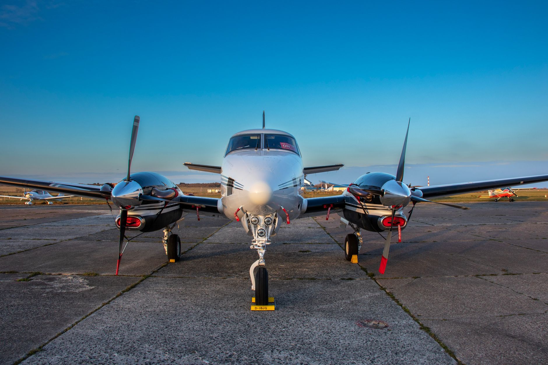 A front view of a twin-engine aircraft on tarmac under clear blue skies.