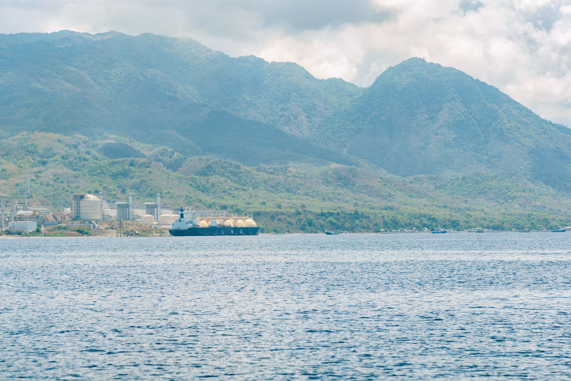 LNG tanker at an industrial port with lush mountains in the background under a cloudy sky.