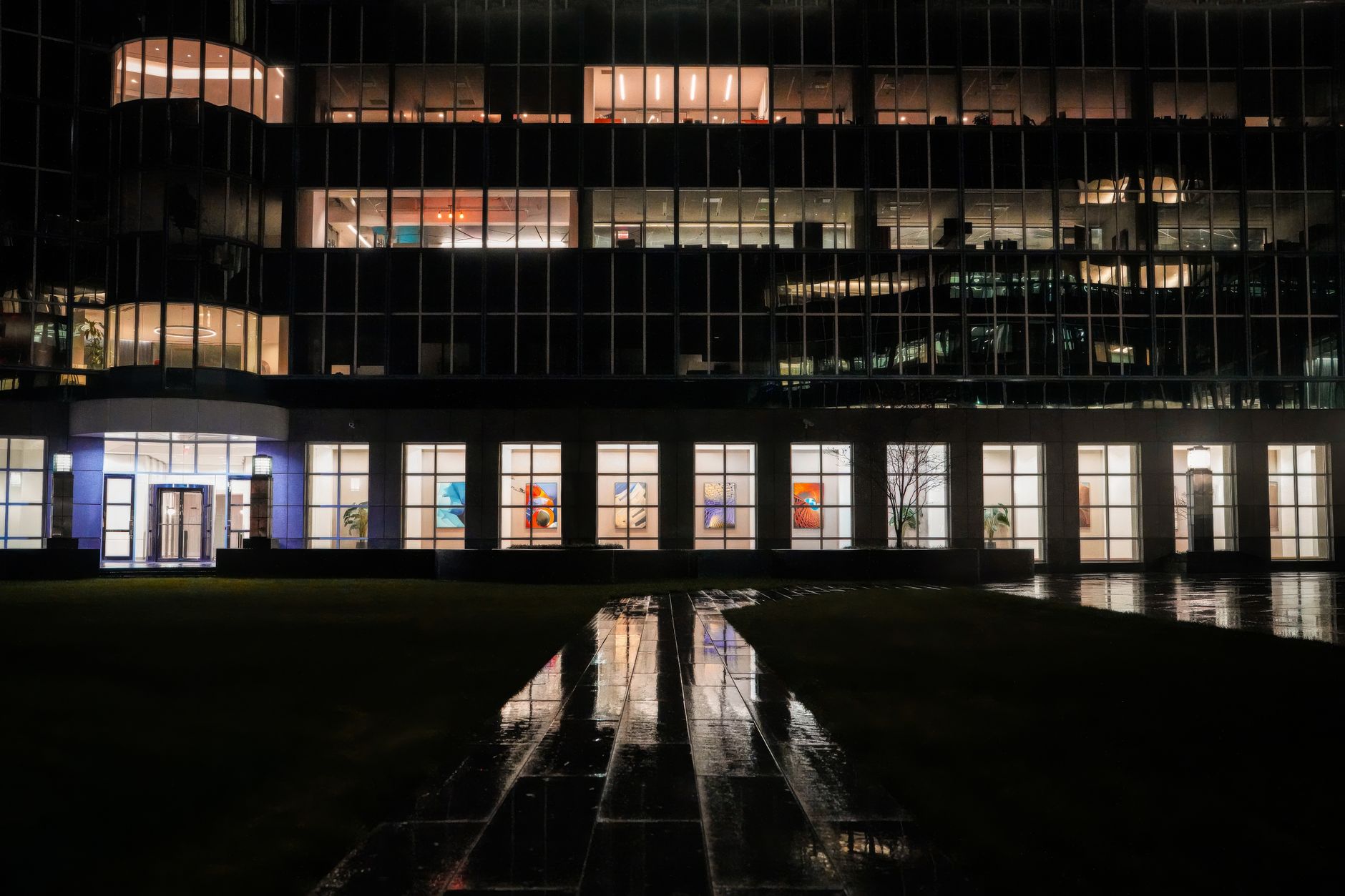 Illuminated office building exterior with reflections on wet pavement at night.